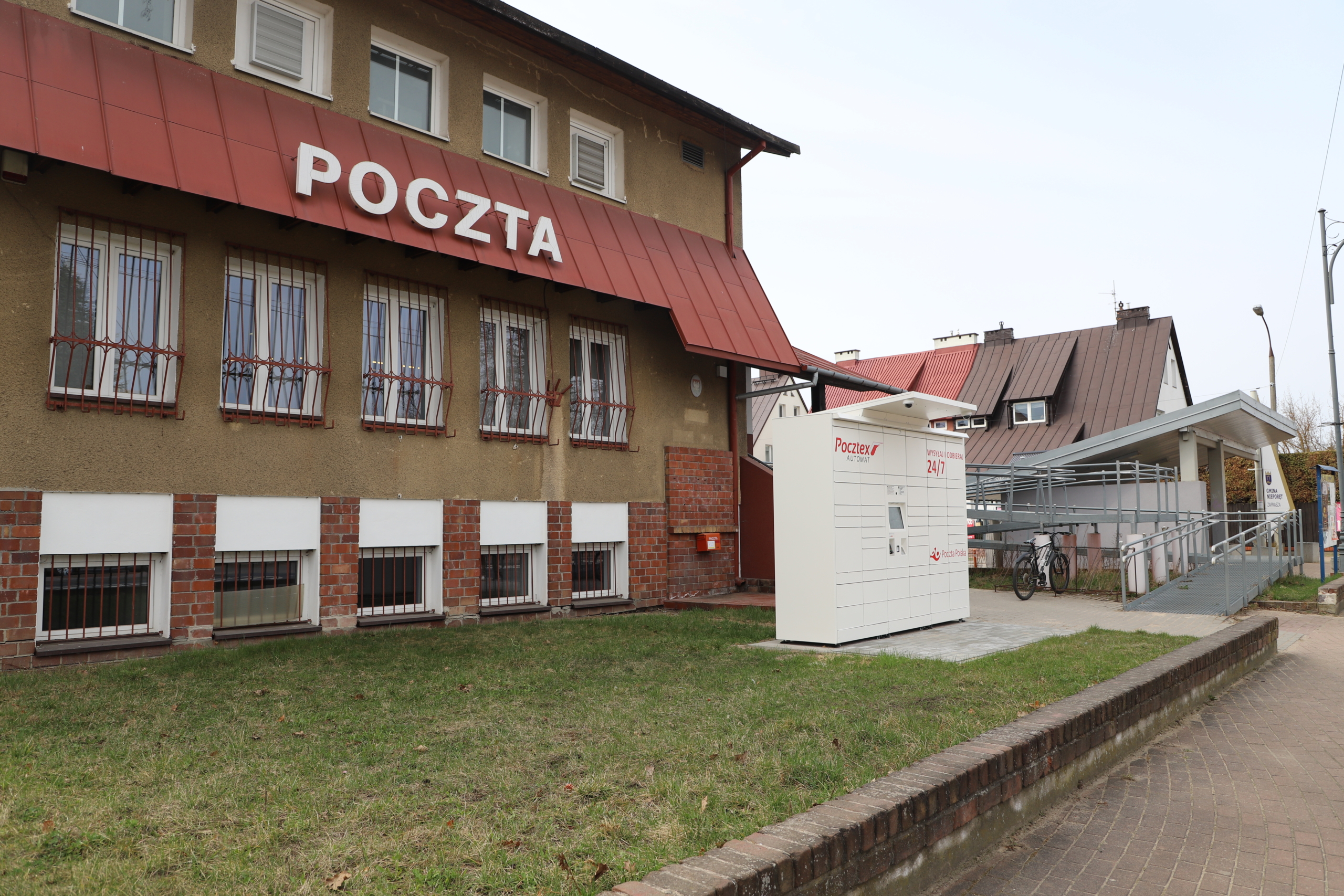 Na zdjęciu budynek Urzędu Pocztowego przy ul. Chłodnej 1 A w Nieporęcie. Z przodu budynku widoczny paczkomat oraz wiata przystanku autobusowego.
